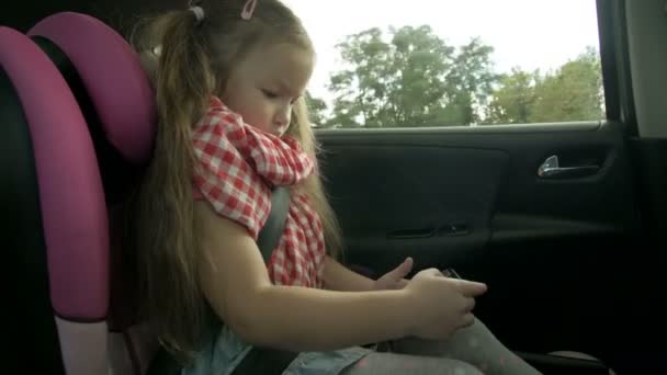 Χαριτωμένο κοριτσάκι απασχολημένος με έξυπνο τηλέφωνο, ενώ κάθεται στο πίσω κάθισμα του αυτοκινήτου. Υπέροχο παιδί στοιχειώδη ηλικία παίζει στο κινητό τηλέφωνο κατά τη διάρκεια του ταξιδιού — Αρχείο Βίντεο