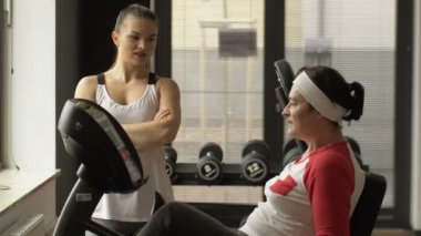 Orta yaşlı bir kadın spor salonunda bisiklet simülatöründe egzersiz yaparken kadın antrenör yanında duruyordu.