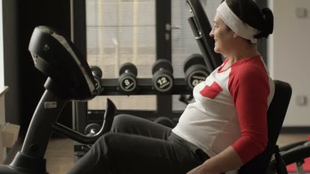 Kobieta w średnim wieku robi ćwiczenia cardio na rowerze papeteryjnym w siłowni lub centrum fitness. — Wideo stockowe