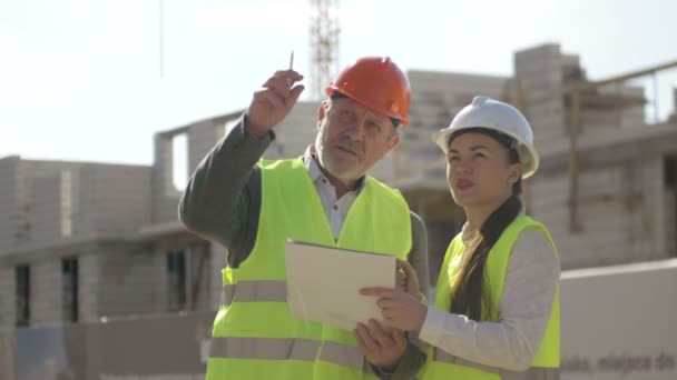 Man and young woman in helmets on the background of a house under construction. — Stock Video