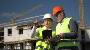 Miğferli bir adam ve genç bir kadın inşaat halindeki bir evin arka planında.