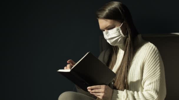 Ragazza con una maschera medica con un diario nero in mano. Legge con attenzione. . — Video Stock