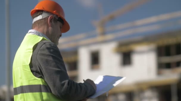 Elderly man in a signal vest and hard hat writes something. Against the background of an object under construction. — Stock Video