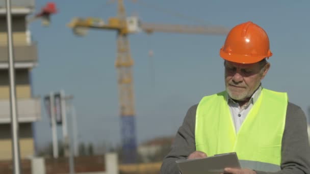 Muž v signální vestě a klobouku pečlivě sleduje, co se děje na staveništi a něco píše.. — Stock video
