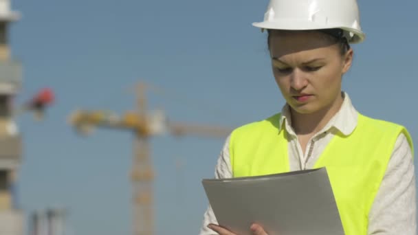 Een bouwvrouw met een helm schrijft iets. Ze is sereen en geconcentreerd. Tegen de achtergrond van de bouwplaats. — Stockvideo