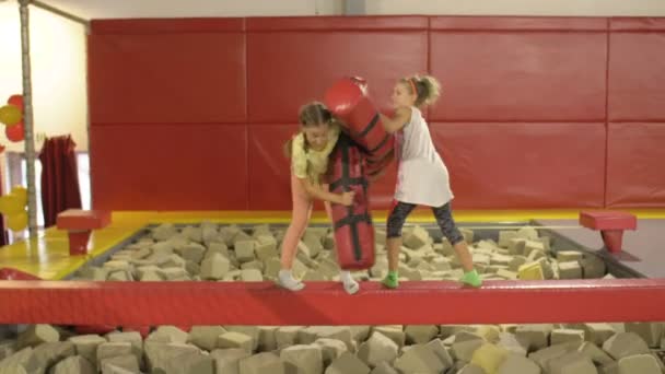 Dos muchachas de la edad media de la escuela se empujan alegremente uno a otro con los rodillos suaves. Gladiadores. Diversión en el parque de trampolín . — Vídeos de Stock