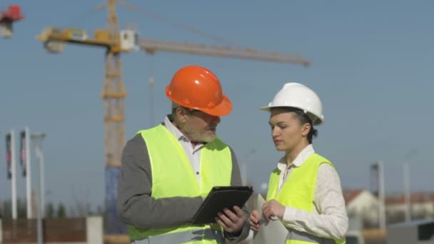 Hombre mayor con una tableta en las manos y una joven en un sitio de construcción. Los constructores discuten temas de trabajo . — Vídeos de Stock