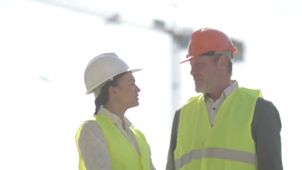 Two builders, a man and a young woman, are discussing work issues. Against the background of a crane. — Stock Video
