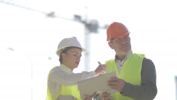 Δύο χτίστες, ένας άνδρας και μια νεαρή γυναίκα, συζητούν θέματα εργασίας. Με φόντο γερανό. — Αρχείο Βίντεο