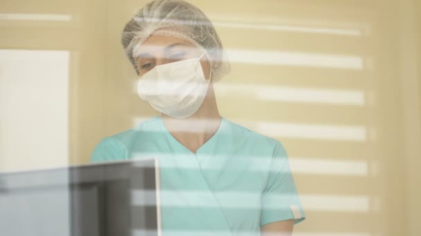 Pielęgniarka lub lekarz w masce medycznej i jednorazowej czapce patrzy na ekran monitora. — Wideo stockowe