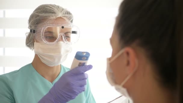 Sjuksköterska i medicinsk mask och glasögon mäter patientens temperatur med en beröringsfri infraröd termometer. — Stockvideo