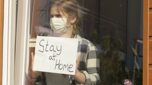 Dziewczyna w masce medycznej stoi przy oknie z plakatem Pobyt w domu. Kwarantanna. — Wideo stockowe