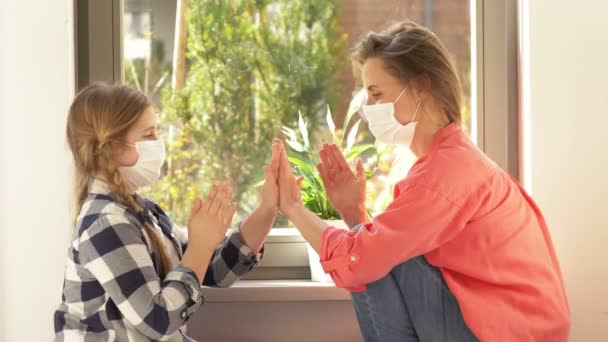 Mamma och dotter hemma under karantänen. Kvinna och flicka i medicinska masker. Mot bakgrund av fönstret. — Stockvideo