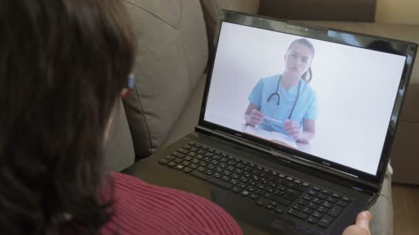 オンライン医師。彼女のアパートでソファの上にラップトップと一緒に座っている女性は、インターネットを介して彼女の医者に相談します. — ストック動画