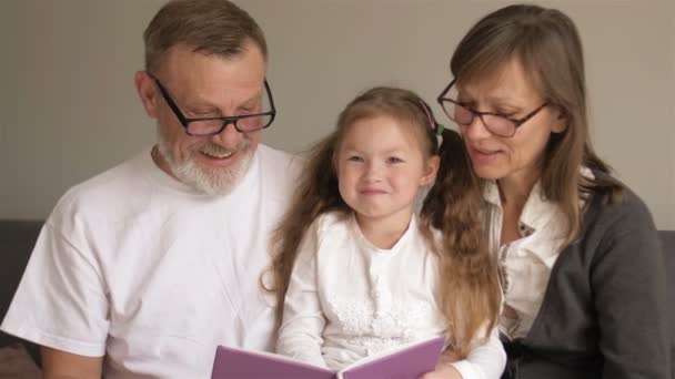 Portrét roztomilých prarodičů s jejich vnoučátko čtení zajímavý příběh z knihy společně, zatímco sedí na pohovce v obývacím pokoji — Stock video