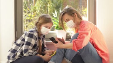 Tıbbi maskeli anne ve kız ellerinde cep telefonlarıyla pencerede oturuyorlar. Karantina sırasında eğlence.