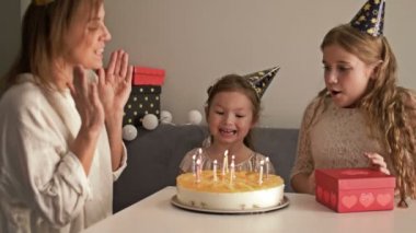 Gülümseyen anne ve kızları evde doğum günü pastası, aile ve kutlama konseptine mum üflüyorlar.