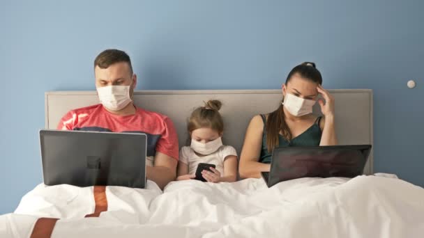 Ehepaar in medizinischen Masken auf einem Bett mit Laptop liegend. Kleines Mädchen mit Handy in der Hand. Pastime während der COVID-19 Coronavirus Epidemie. — Stockvideo