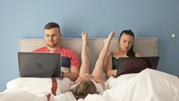 Online werk thuis. Jong getrouwd stel dat met laptops werkt terwijl ze in bed liggen. Klein meisje is erg verveeld, ze heeft ouderlijke aandacht nodig. — Stockvideo