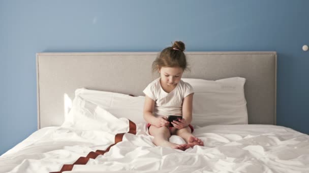 Klein meisje zit op het bed met een mobiele telefoon. Hobbi van moderne kinderen. — Stockvideo