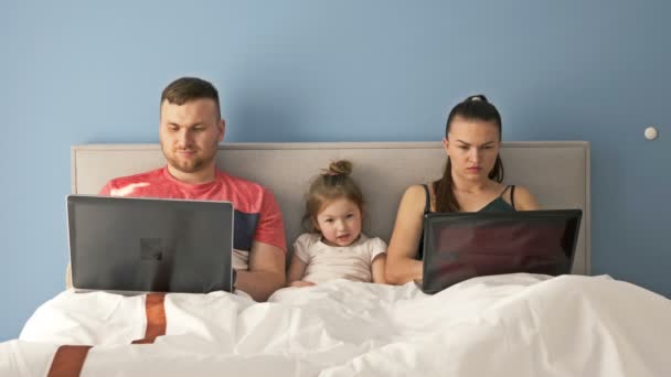 Fiatal házaspár, akik laptoppal dolgoznak, miközben ágyban fekszenek. A kislány nagyon unatkozik, szülői felügyeletet igényel. A szülők megértik a babát.. — Stock videók