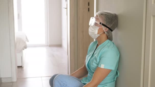 Erschöpft setzt sich die Krankenschwester in Schutzbrille und medizinischer Maske auf den Fußboden im Krankenhausflur. Sie zieht ihre Latexhandschuhe aus. — Stockvideo