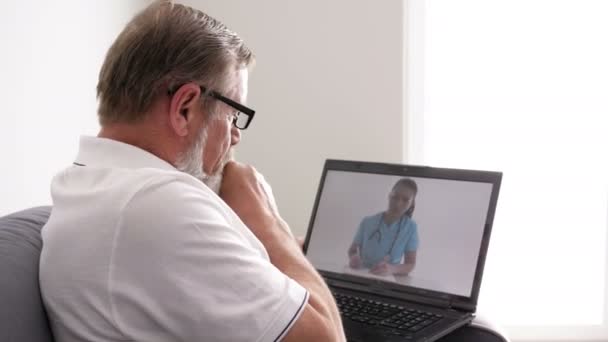Telemedycyna. Starszy mężczyzna siedzący z laptopem na kanapie w jej mieszkaniu konsultuje się z lekarzem przez Internet. — Wideo stockowe