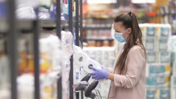 Donna carina che indossa una maschera protettiva e guanti vicino agli scaffali con carta igienica in un supermercato durante un'epidemia di virus . — Video Stock