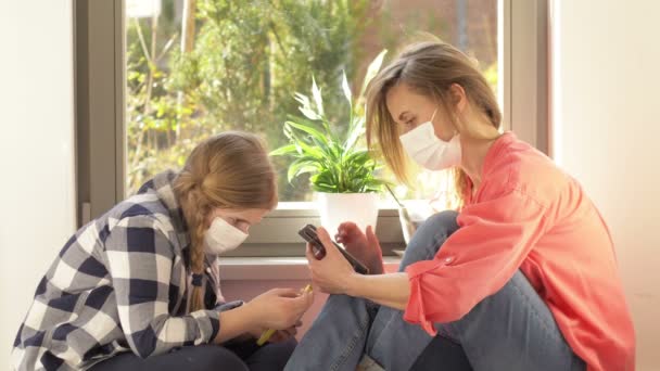 Mamma e figlia in maschere mediche sono seduti alla finestra con i telefoni cellulari. Passatempo durante la quarantena . — Video Stock