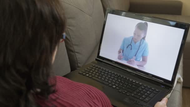 Çevrimiçi doktor. Evindeki kanepede dizüstü bilgisayarla oturan bir kadın doktoruna internet üzerinden danışıyor.. — Stok video