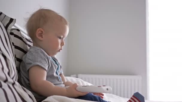 Bebê bonito está sentado na cama e examina com interesse a meia em sua perna . — Vídeo de Stock