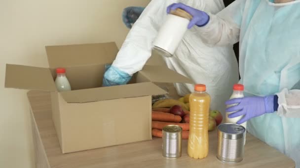 Koruyucu eldivenli iki gönüllü yiyecek ve gerekli şeyleri karton bir kutuya koydu. Koronavirüs salgınında ihtiyacı olanlara yardım ediyor.. — Stok video