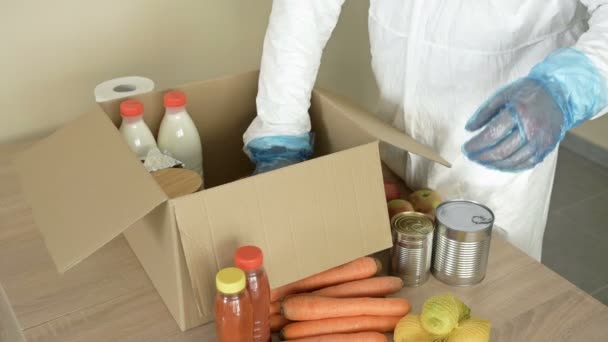 Εθελοντής με προστατευτική ενδυμασία βάζει φαγητό σε ένα κουτί. Για άτομα σε δύσκολες καταστάσεις λόγω της επιδημίας covid-19. — Αρχείο Βίντεο