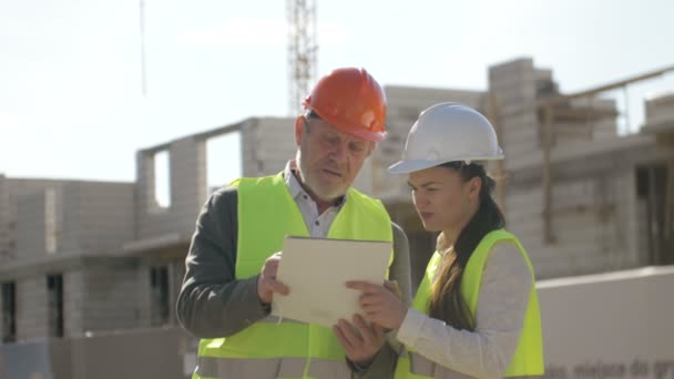 Man en jonge vrouw in helmen op de achtergrond van een huis in aanbouw. — Stockvideo