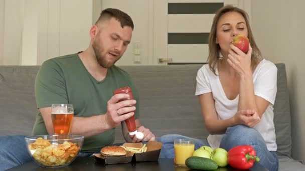 Dieta saludable y poco saludable. La pareja joven tiene diferentes hábitos alimenticios. El marido está feliz de comer comida rápida, la esposa es vegetariana . — Vídeo de stock