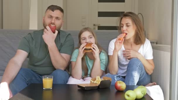 Una giovane famiglia di tre persone sta cenando. Papà e mamma mangiano cibo vegetariano. Adolescente ragazza piace fast food. Dieta sana e malsana . — Video Stock