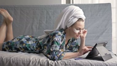 Duştan sonra çekici bir ev hanımı kanepede yatarken online mağazadan alışveriş yapmaktan mutluluk duyar. Kadın dizüstü bilgisayar ve ödeme kartı kullanıyor..