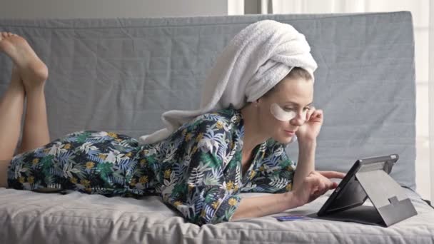 Atractiva ama de casa después de una ducha está feliz de comprar en la tienda en línea mientras está acostado en el sofá. Mujer utiliza un ordenador portátil y una tarjeta de pago . — Vídeos de Stock