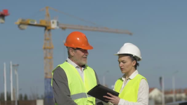 Homem idoso e uma jovem mulher com um tablet nas mãos em um canteiro de obras. Construtores discutir questões de trabalho . — Vídeo de Stock