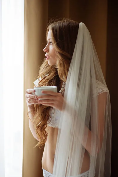 Bruden i sexuella underkläder med kopp kaffe nära windiw av lägenhet på röda hotellbyggnaden. — Stockfoto
