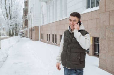 Açık kış portre yakışıklı delikanlı ile belgili tanımlık telefon için. Güzel genç onun ceket ve üzerinde bir şehir sokak, köknar ağaçlarının arka plan poz yelek.