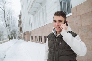 Açık kış portre yakışıklı delikanlı ile belgili tanımlık telefon için. Güzel genç onun ceket ve üzerinde bir şehir sokak, köknar ağaçlarının arka plan poz yelek.