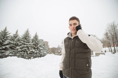 Açık kış portre yakışıklı delikanlı ile belgili tanımlık telefon için. Güzel genç onun ceket ve üzerinde bir şehir sokak, köknar ağaçlarının arka plan poz yelek.