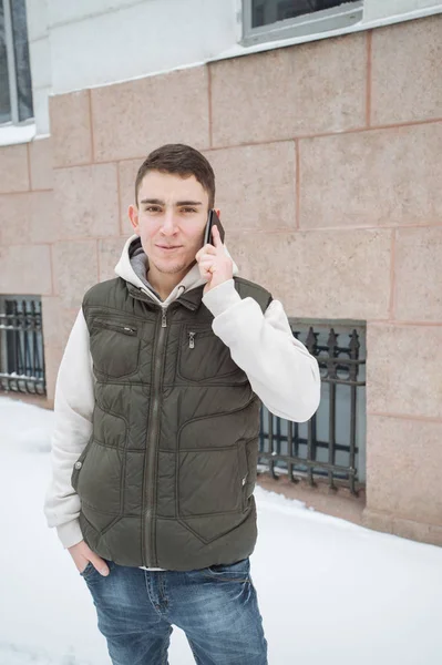 Utomhus vinter porträtt för ung stilig man med telefonen. Vackra tonåring i hans jacka och väst poserar på en stadsgata, bakgrund av granar. — Stockfoto