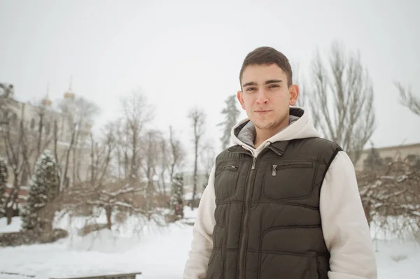 Winterporträt für einen jungen, gut aussehenden Mann. schöner Teenager in Jacke und Weste posiert auf einer Straße in der Stadt, Hintergrund von Tannen. — Stockfoto