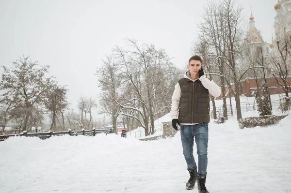 Kültéri téli portré jóképű fiatalember a telefon. A kabát és mellény pózol egy városi utca, háttérben a fenyők a szép kamasz. — Stock Fotó