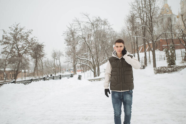 Outdoor winter portrait for young handsome man with the phone. Beautiful teenager in his jacket and vest posing on a city street, background of fir trees.