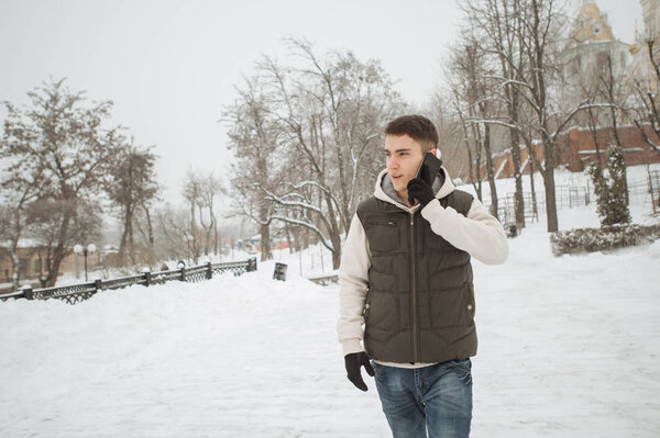 Outdoor winter portrait for young handsome man with the phone. Beautiful teenager in his jacket and vest posing on a city street, background of fir trees.