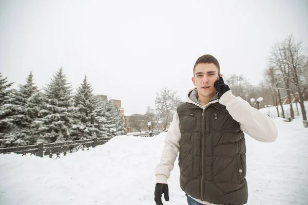 Outdoor winter portrait for young handsome man with the phone. Beautiful teenager in his jacket and vest posing on a city street, background of fir trees. — Stock Photo, Image