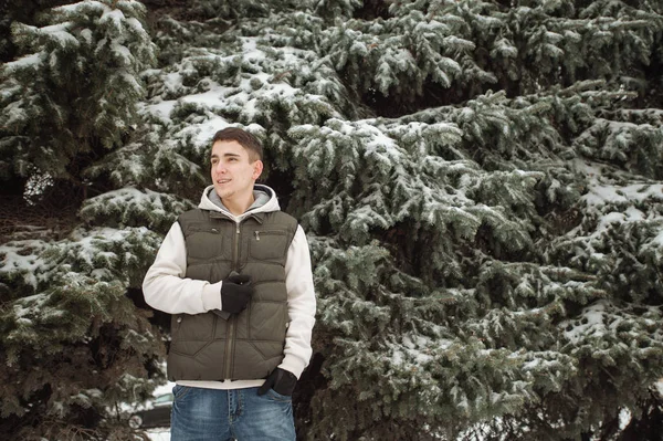 Utomhus vinter porträtt för ung stilig man. Vackra tonåring i hans jacka och väst poserar på en stadsgata, bakgrund av granar. — Stockfoto